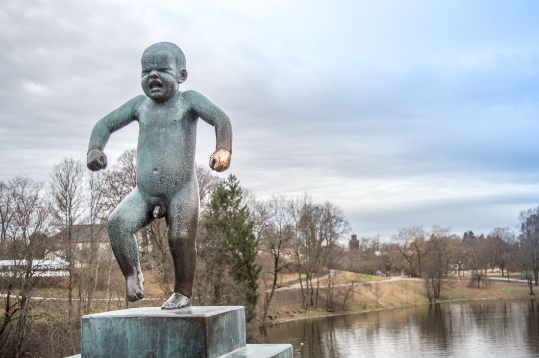 One Day in Oslo: This is a picture of the Angry Boy statue. His hand shimmers golden and in the background there is a body of water with sparse vegetation to either side.