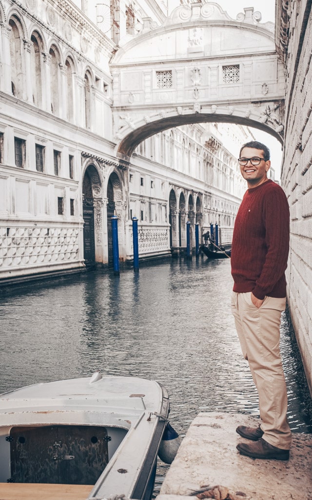 Man posing for a photo on a ledge next to a canal in front of the famous Bridge of Sighs in Venice, Italy.