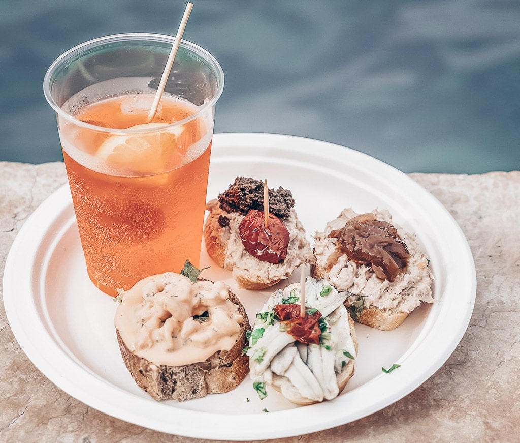 Traditional cicchetti on a plate along with a glass of Aperol Spritz next to a canal in Venice.