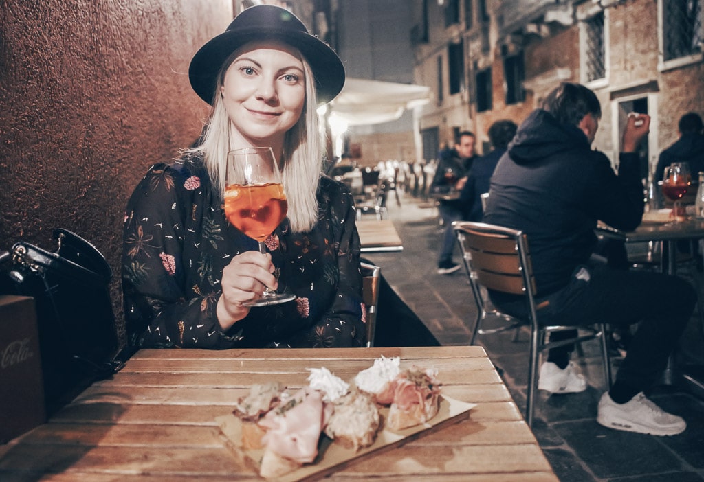 Venice cicchetti: Beautiful woman holding a glass of Aperol Spritz seated at a table containing a plate of cicchetti.