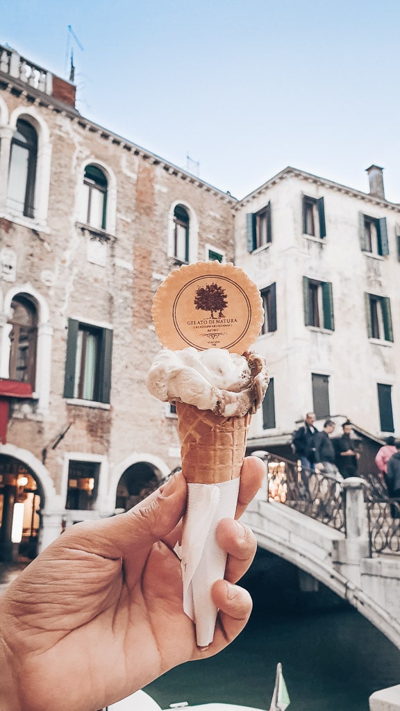 Man holding a cone of pistachio gelato (Italian ice cream) near a canal and bridge in Venice.